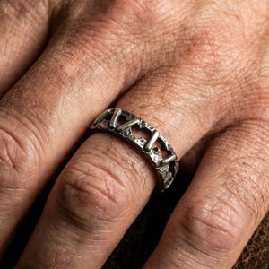 Op de afbeelding: Een zilverkleurige ring met een donkere, geoxideerde afwerking en een getextureerd, ruw oppervlak. De band heeft een herhalend patroon van verticale staven gescheiden door open, onregelmatige uitsparingen, wat zorgt voor een uitgesproken, verweerde en industri&euml;le uitstraling.