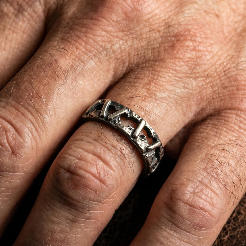 Op de afbeelding: Een zilverkleurige ring met een donkere, geoxideerde afwerking en een getextureerd, ruw oppervlak. De band heeft een herhalend patroon van verticale staven gescheiden door open, onregelmatige uitsparingen, wat zorgt voor een uitgesproken, verweerde en industri&euml;le uitstraling.