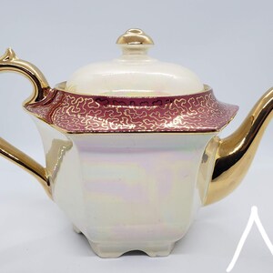 Peut inclure: Une théière en porcelaine blanche avec une anse et un bec verseur dorés. La théière a un corps carré avec une bande rouge et dorée autour du haut. Le couvercle est blanc avec un bouton doré.