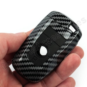 May include: A black and grey carbon fibre patterned car key fob cover. The cover has a rectangular shape with rounded edges and features buttons and a keyhole. The text "CarStyle Accessories" is visible.