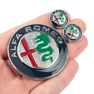 May include: Three silver Alfa Romeo car badges. The largest badge is a round, blue and silver badge with the words "Alfa Romeo" in silver lettering around the outside of the badge. The badge features a red cross and a green snake. The two smaller badges are round and silver with the words "Alfa Romeo" in silver lettering around the outside of the badge. The smaller badges feature a red cross and a green snake.