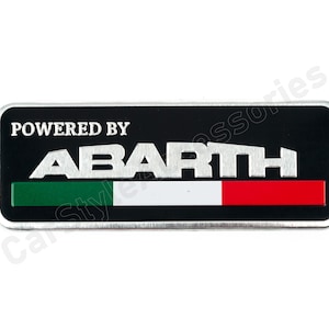Op de afbeelding: Een rechthoekig zwart en zilverkleurig embleem met de tekst "POWERED BY ABARTH" in het wit. Daaronder een horizontale streep in de kleuren van de Italiaanse vlag: groen, wit en rood. Het embleem heeft een zilveren rand.