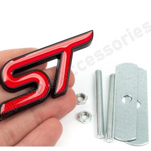 Op de afbeelding: Een rood en zwart ST-embleem met montagemateriaal. Het embleem is glanzend en heeft de letters "ST" in een vetgedrukte, gestileerde lettertype. Inclusief twee bouten, twee moeren en twee metalen montageplaten.