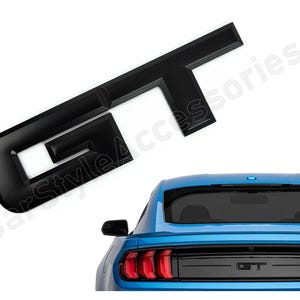 Könnte beinhalten: Schwarzes Plastik-Emblem "GT" für ein Auto. Das Emblem ist auf einem blauen Auto mit schwarzer Heckscheibe und einem schwarzen "GT"-Emblem am Heckstoßfänger zu sehen.