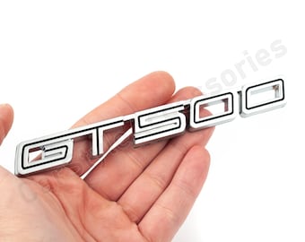 May include: Silver GT500 emblem with black accents. The emblem is held in a hand against a white background. The letters and numbers are outlined in black.