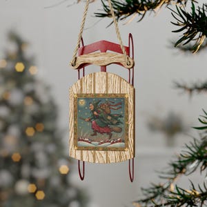 Puede incluir: Adorno navideño de trineo de madera con marco rojo y patines metálicos. El trineo presenta una ilustración enmarcada de una bruja volando en una escoba sobre casas cubiertas de nieve. El adorno cuelga de una cuerda de hilo natural.