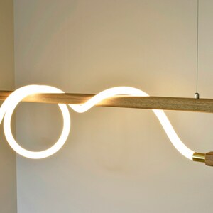 May include: A modern pendant light fitting with a wooden frame and a white LED light that is shaped like a wave. The light is suspended from the ceiling by a thin wire.