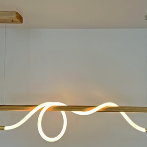 May include: A modern, minimalist pendant light fitting with a wooden frame and a white LED light that is shaped like a wave. The light is suspended from the ceiling by two wires.