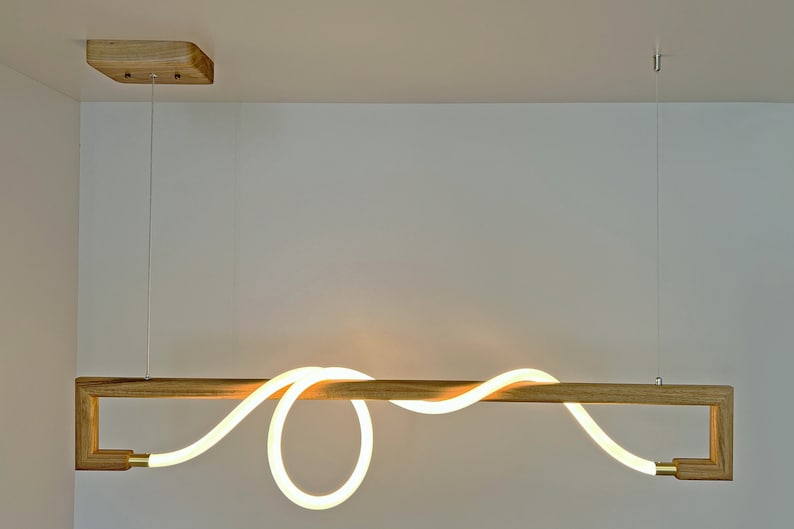May include: A modern, minimalist pendant light fitting with a wooden frame and a white LED light that is shaped like a wave. The light is suspended from the ceiling by two wires.