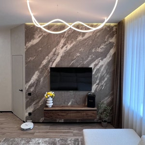 May include: A modern living room with a white, wavy light fixture hanging from the ceiling. The room features a large, flat-screen TV mounted on a wall with a marble-like finish. A wooden entertainment center with drawers sits below the TV. A white couch with a textured fabric sits in the foreground.