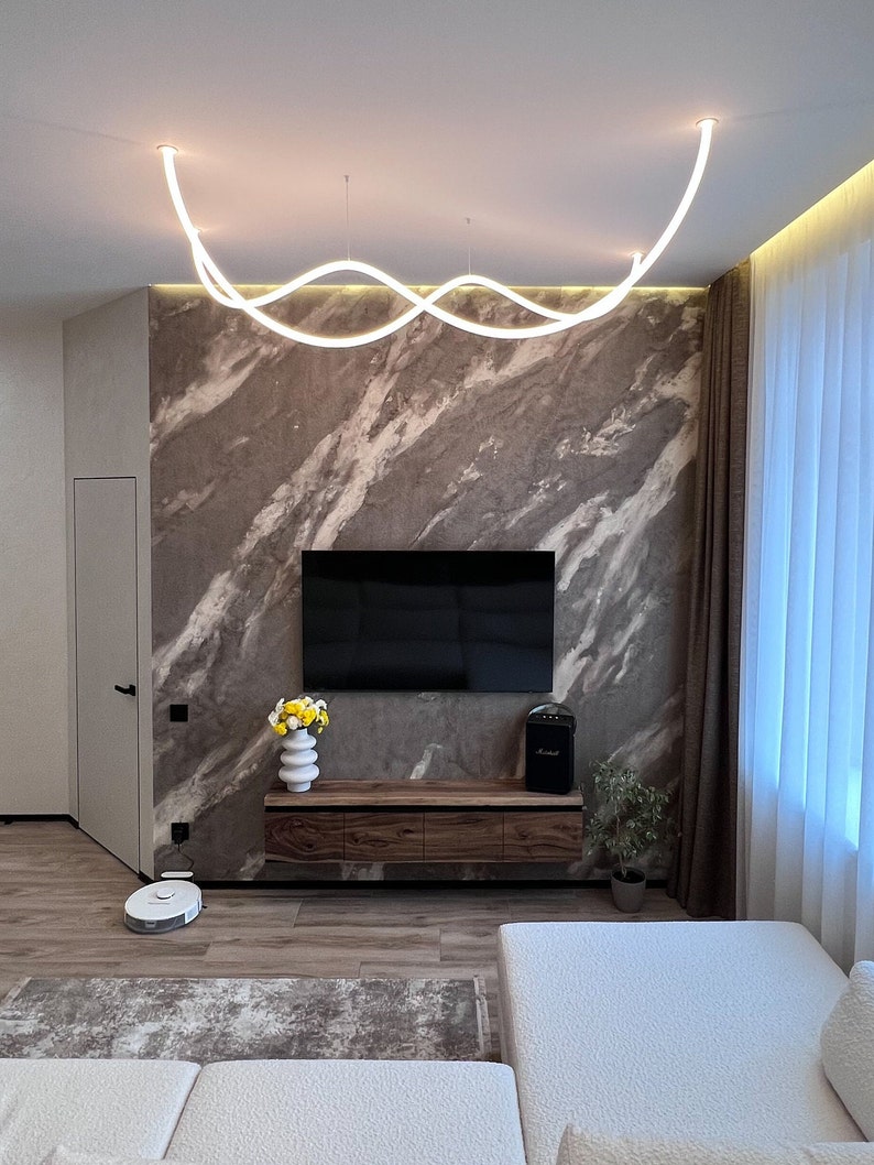 May include: A modern living room with a white, wavy light fixture hanging from the ceiling. The room features a large, flat-screen TV mounted on a wall with a marble-like finish. A wooden entertainment center with drawers sits below the TV. A white couch with a textured fabric sits in the foreground.