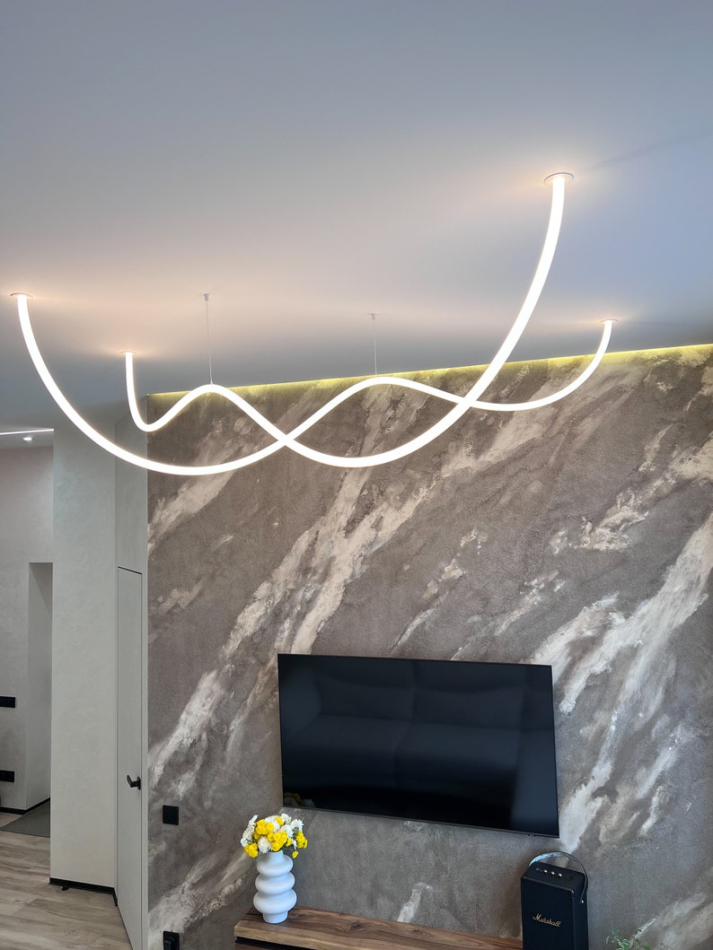 May include: A modern, white, curved light fixture hangs from the ceiling in a room with a gray and white marble wall. The light fixture is made of three separate pieces that are connected in a wave-like pattern. A television is mounted on the wall below the light fixture.