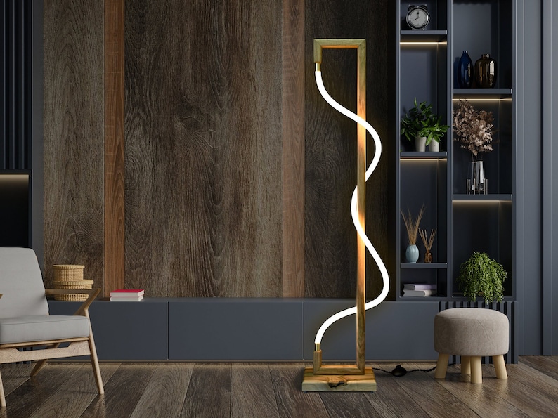 Pu&ograve; includere: Una lampada da terra moderna con una struttura in metallo dorato e una luce LED bianca che si curva a forma di onda. La lampada &egrave; posizionata su un pavimento in legno in una stanza con una parete in legno e un mobile blu scuro.