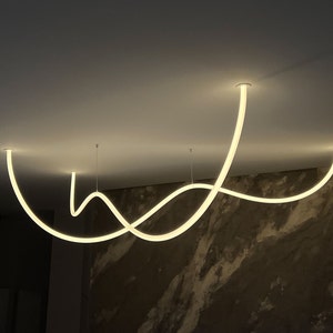 May include: A modern, white, curved light fixture hangs from the ceiling. The fixture is made of a thin, flexible material and is shaped like a series of waves. The light fixture is illuminated, casting a warm glow on the surrounding area.