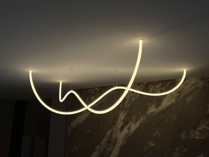 May include: A modern, white, curved light fixture hangs from the ceiling. The fixture is made of a thin, flexible material and is shaped like a series of waves. The light fixture is illuminated, casting a warm glow on the surrounding area.