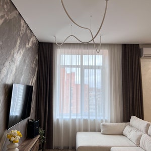 May include: A white sectional sofa with a white throw pillow sits in a living room with brown curtains, a large window, and a white ceiling with a unique white light fixture.