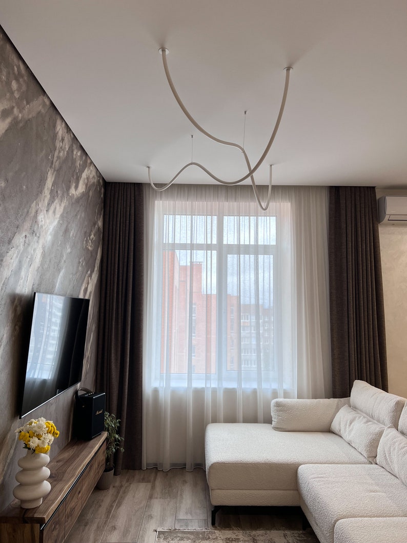 May include: A white sectional sofa with a white throw pillow sits in a living room with brown curtains, a large window, and a white ceiling with a unique white light fixture.