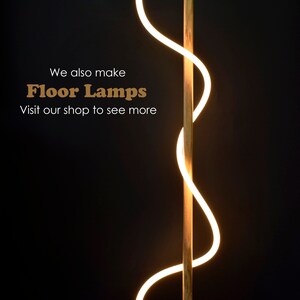 May include: A modern floor lamp with a wooden base and a curved, white LED light. The lamp is lit and casts a warm glow. The text "We also make Floor Lamps Visit our shop to see more" is visible in the image.