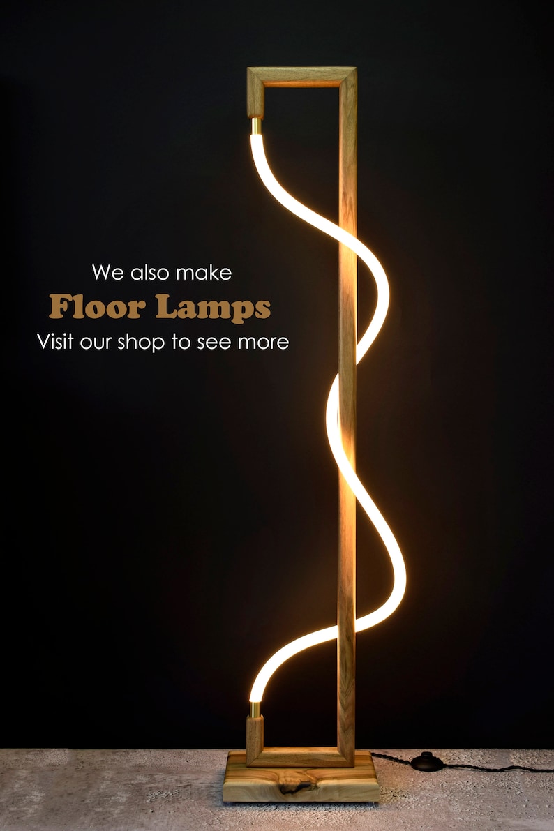 May include: A modern floor lamp with a wooden base and a curved, white LED light. The lamp is lit and casts a warm glow. The text "We also make Floor Lamps Visit our shop to see more" is visible in the image.