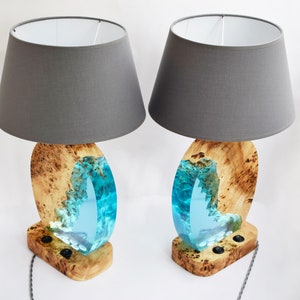 Puede incluir: Dos l&aacute;mparas de mesa a juego con pantallas de tela gris. Las bases son de madera con una incrustaci&oacute;n de resina azul que se asemeja a una ola.