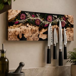 May include: A rectangular magnetic knife holder featuring a live-edge burl wood section and a black resin inlay embedded with dried pink flowers and white grasses. Four stainless steel kitchen knives with black handles are attached to the surface.