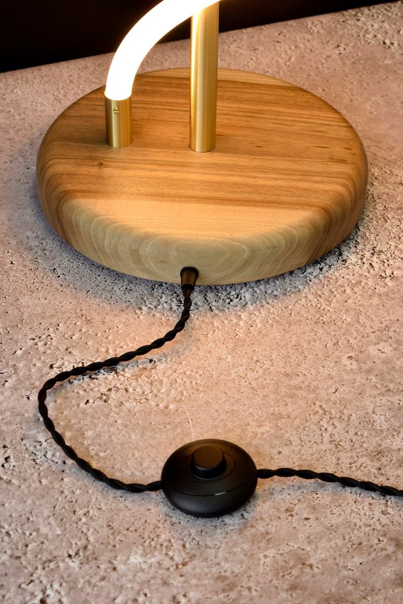 May include: A wooden table lamp with a gold metal base and a white curved light fixture. The lamp has a black cord with a foot switch.