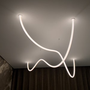 May include: A modern ceiling light fixture with three white, curved, LED light tubes. The light tubes are suspended from the ceiling and create a unique, abstract design.