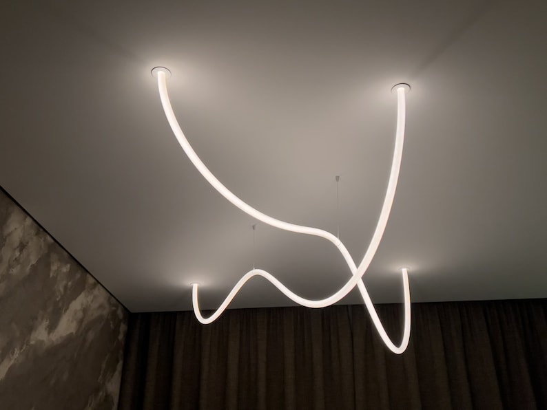 May include: A modern ceiling light fixture with three white, curved, LED light tubes. The light tubes are suspended from the ceiling and create a unique, abstract design.