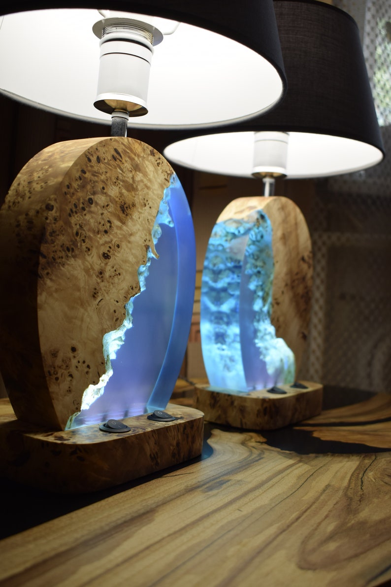 Puede incluir: Dos l&aacute;mparas de mesa de madera con incrustaciones de resina azul y pantallas de l&aacute;mpara blancas y negras. Las l&aacute;mparas tienen un acabado de madera natural y est&aacute;n iluminadas con luz blanca c&aacute;lida.