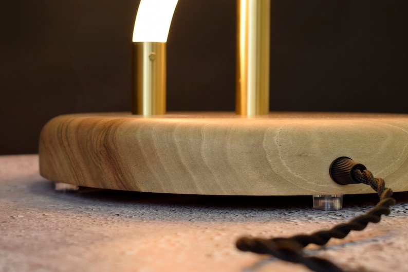 May include: A wooden table lamp with a gold metal base and a curved white light fixture. The lamp has a black cord and a clear plug.