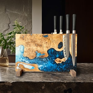 Könnte beinhalten: Ein Messerblock aus Holz mit einem einzigartigen Design. Der Block besteht aus einer Kombination aus hellbraunem Holz und leuchtend blauer Kunstharz, mit vier eingesetzten Messern. Der Block wird von zwei kleinen Holzfüßen getragen, und die Messer haben schwarze Griffe.