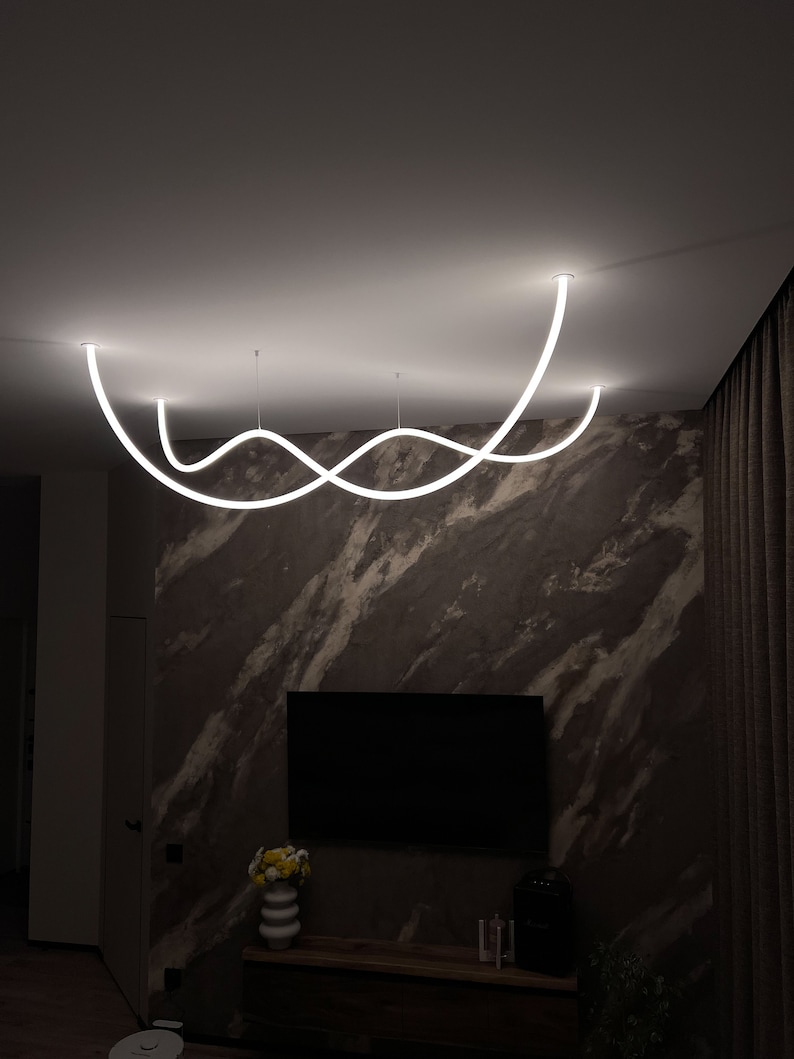 May include: A modern white LED light fixture with a curved design hangs from the ceiling in a living room. The light fixture is suspended over a dark brown marble wall with a flat screen TV mounted on the wall.