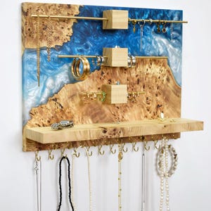 Puede incluir: Organizador de joyas de madera con un diseño de borde vivo, con un río de resina azul. Tiene barras y ganchos de latón para colgar collares, pendientes y pulseras. El organizador incluye un estante.