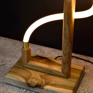 Pu&ograve; includere: Una lampada da terra in legno con una luce LED bianca curva. La lampada ha una finitura in legno naturale e una base rettangolare.