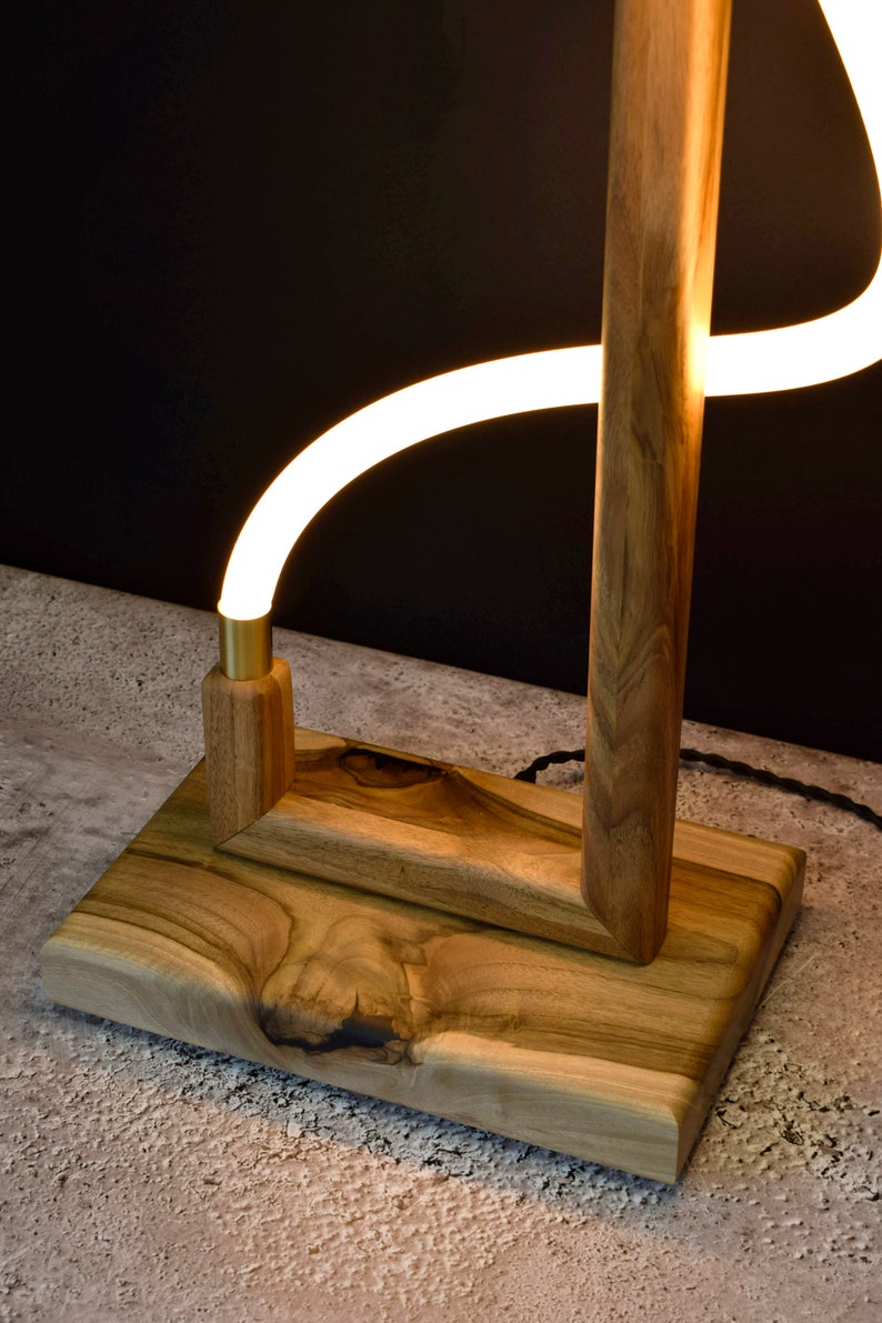 Pu&ograve; includere: Una lampada da terra in legno con una luce LED bianca curva. La lampada ha una finitura in legno naturale e una base rettangolare.