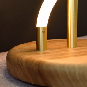 May include: A modern table lamp with a curved, white light source and a gold metal base. The base is mounted on a round, wooden platform.