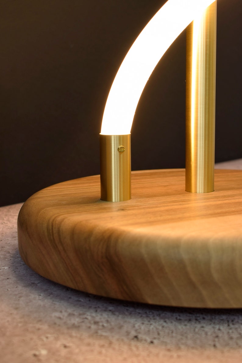 May include: A modern table lamp with a curved, white light source and a gold metal base. The base is mounted on a round, wooden platform.