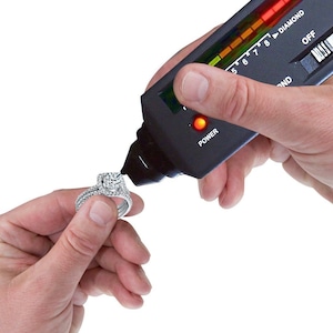 May include: A black diamond tester with a red light and a scale from 5 to 8. The tester is being used to test a diamond ring.