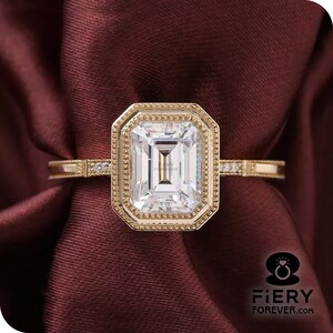 May include: A gold engagement ring with a large emerald cut diamond set in a bezel setting with a decorative border. The ring has a thin band with small diamonds on each side.