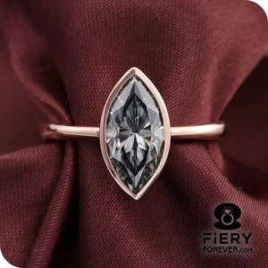 Puede incluir: Un anillo de compromiso de oro rosa con un diamante de talla marquesa. El diamante está engastado en garras y tiene un engaste de bisel alrededor del borde. El anillo está sobre un fondo de tela roja.