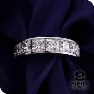 May include: A silver band ring with a row of square-shaped clear gemstones. The ring is on a dark blue fabric background.