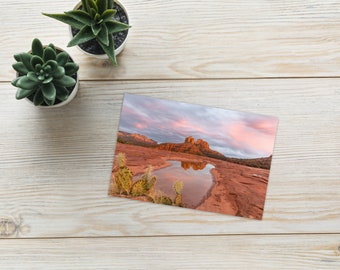 Op de afbeelding: Een ansichtkaart met een schilderachtig landschap met rode rotsformaties die worden weerspiegeld in een plas water. De lucht is helder roze en oranje, wat een prachtige zonsondergang creëert.