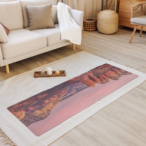 Può includere: Un tappetino da yoga lungo e rettangolare con un'immagine panoramica di una catena montuosa al tramonto. Il tappetino è rosa e arancione con formazioni rocciose marroni. Il tappetino è sdraiato su un tappeto bianco in un soggiorno.