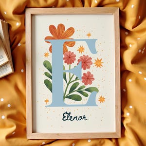 Op de afbeelding: Een ingelijste kunstafdruk met een bloemenontwerp en de letter "E" in lichtblauw. Het kunstwerk bevat oranje en rode bloemen, groene bladeren en de naam "Elenor" in script. De lijst is van lichtgekleurd hout.