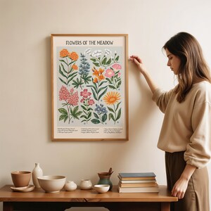 K&ouml;nnte beinhalten: Ein botanisches Illustrationsposter im Vintage-Stil mit dem Titel "Flowers of the Meadow", das eine Vielzahl von bunten Blumen zeigt, deren Namen und lateinische Bezeichnungen darunter aufgelistet sind. Das Poster ist in einem hellen Holzrahmen gerahmt und h&auml;ngt an einer wei&szlig;en Wand.