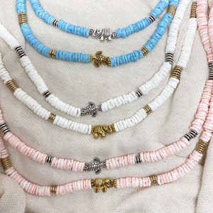 May include: A collection of beaded necklaces in shades of blue, white, and pink. Each necklace features small, flat beads and decorative metal charms, including elephants and turtles. The necklaces are arranged on a neutral fabric background.