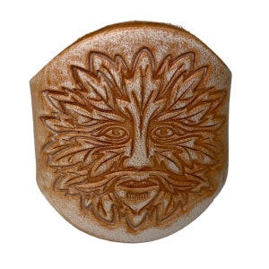 May include: A tan leather cuff bracelet with a carved Green Man face design. The face features detailed eyes, a beard, and a leafy crown. The bracelet has a textured, aged appearance and is set against a white background.