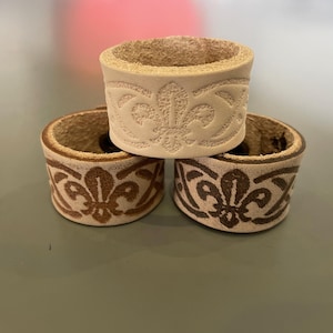 May include: Three stacked, cylindrical leather napkin rings. The rings are beige with a brown fleur-de-lis pattern embossed around the exterior. The interior of the rings is a lighter beige colour.