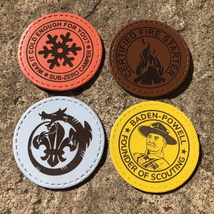 May include: Four circular leather patches with stitched edges. One is orange with a snowflake and text, another is brown with a campfire graphic, a third is light blue with a dragon and fleur-de-lis, and the last is yellow with a portrait and text.