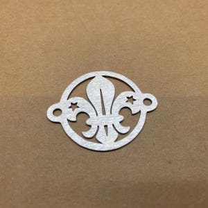 May include: A silver metal charm with a fleur-de-lis design inside a circle, with two stars and two holes for attachment. The charm has a brushed metal finish. Ideal for jewellery making.
